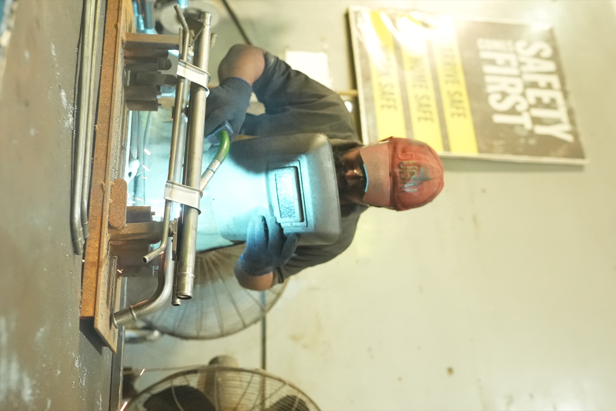 Welder behind a hand shield MIG welding a pipe assembly held in a fixture