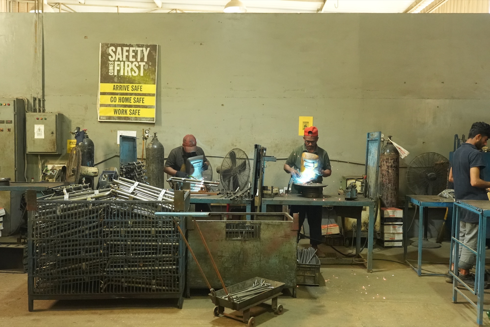Two welders at their stations beneath a Safety Comes First poster, with bins of formed pipe parts in front